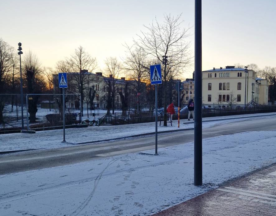 Valokuva. Talvinen maisema suojatiestä, taustalla näkyy vaaleankeltainen koulurakennus.