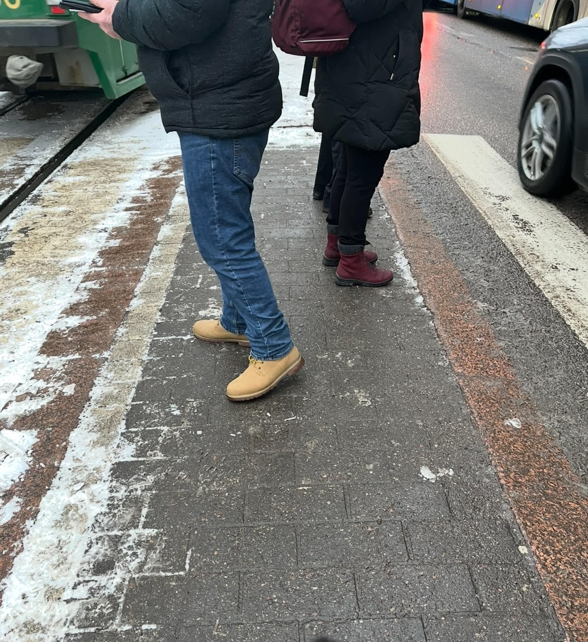 Kuvattu alaviistoon ja kolmen ihmisen jalat näkyvät kapealla saarekkeella. Saarekkeen toiselta puolelta huristaa ratikka ja toiselta auto.